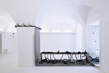 The exhibition Mulberry Tubes by jewellery artist Michaela Rapp shows an installation of havy and small tubes on a low bench on a grey paper that is placed on the wall and the floor; cocoon necklaces inside a see-through net hanging down from the ceiling.