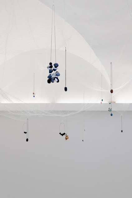 Installation mit farbigen Kokon-Halsketten, die in einem durchsichtigen Netz, das von der Decke herabhängt, platziert sind, als würden sie schweben.