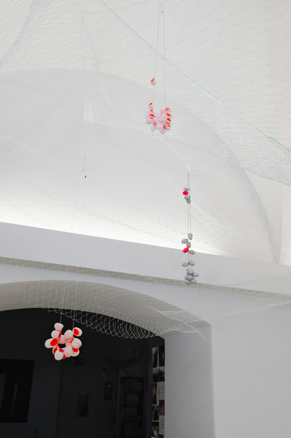 Installation mit farbigen Kokon-Halsketten, die in einem durchsichtigen Netz, das von der Decke herabhängt, platziert sind, als würden sie schweben.