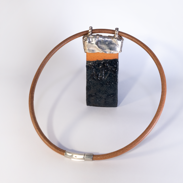 Pendant made of a rectangular black glazed ceramic piece with a silver cap, part of the ceramic is natural reddish, it hangs on a heavy reddish leather strap; the glaze "Fat Lava" looks like the surface of the moon