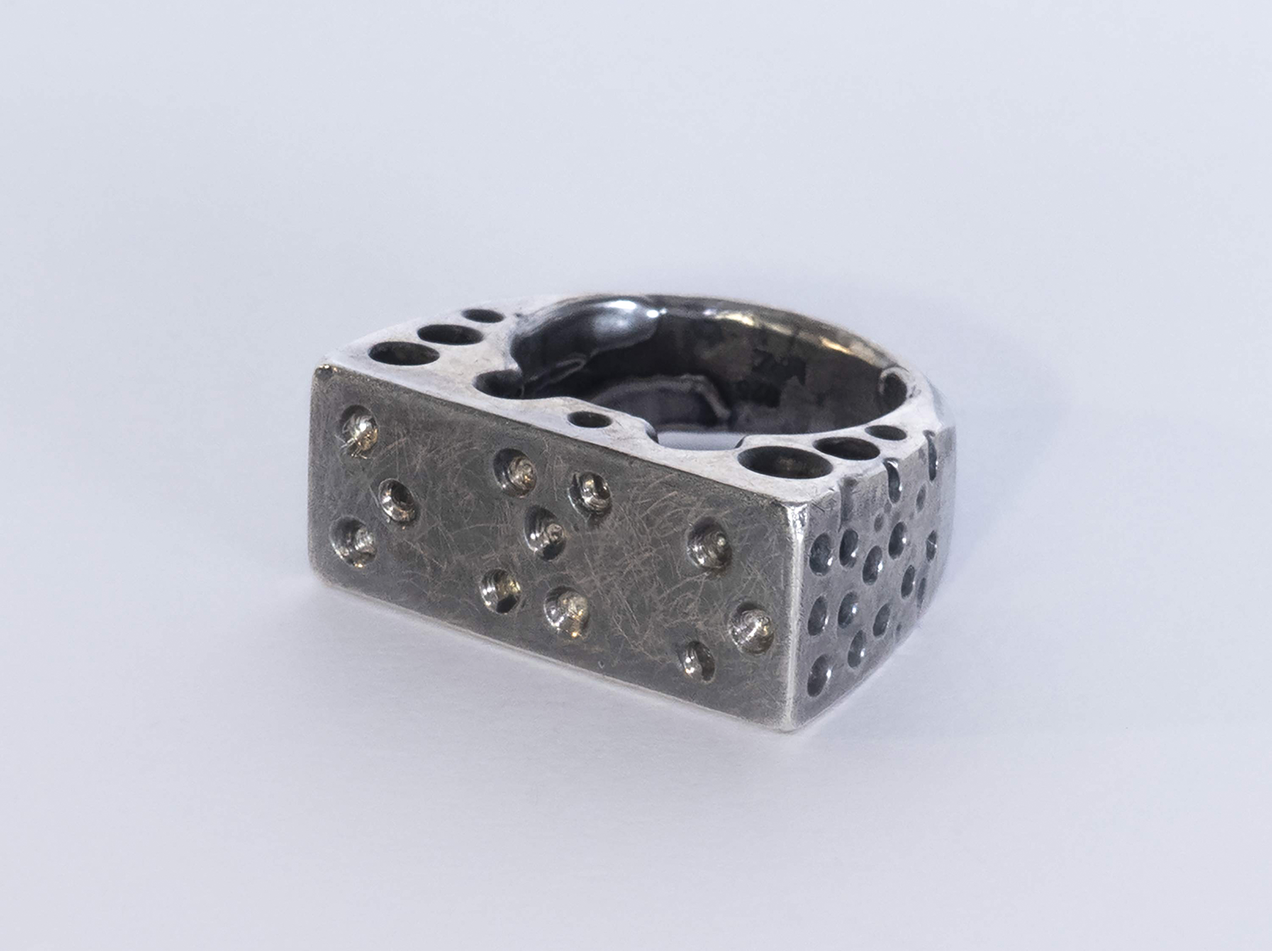 Silver ring showing dimples in braille on the upper surface, the other parts of the ring are perforated