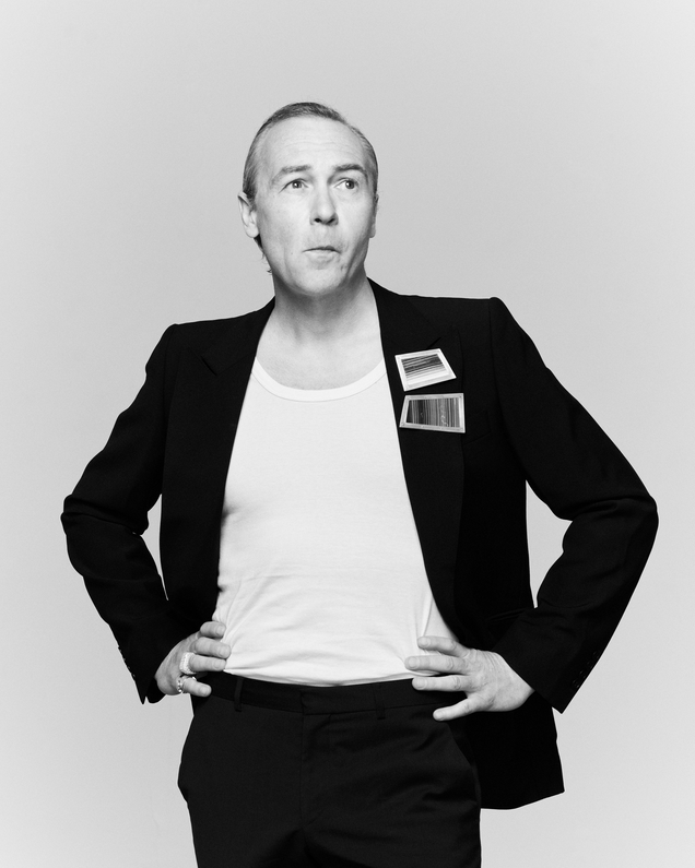 Künstler Robert Jelinek trägt zwei Broschen am Revers des schwarzen Sakkos, sein Haar ist streng zurück gekämmt, er schaut fragend, als hätte er etwas nicht verstanden