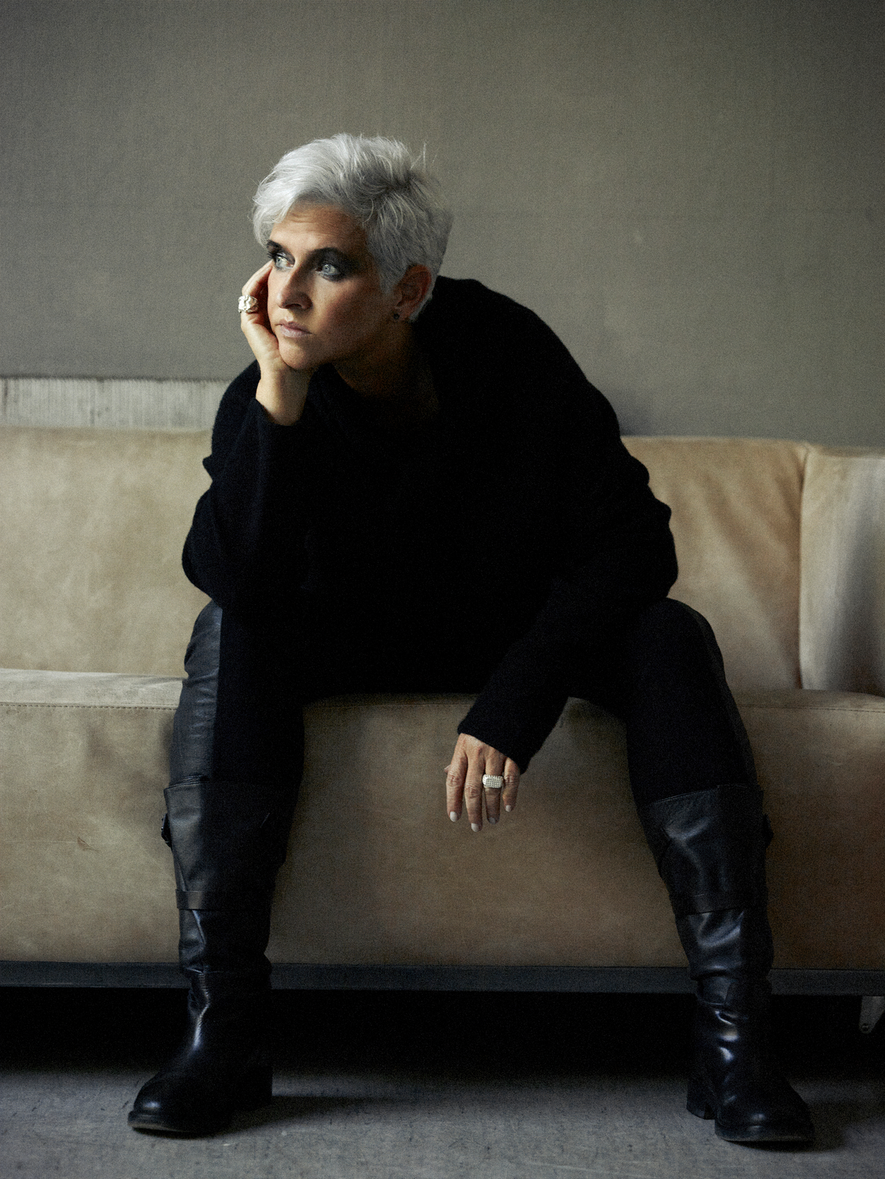 Frau mit silberweißem Haar, schwarzem Pullover, schwarzer Hose und schwarzen Stiefeln, auf einem Sofa sitzend, sie schaut nach links, den Kopf entspannt in die Hand gestützt