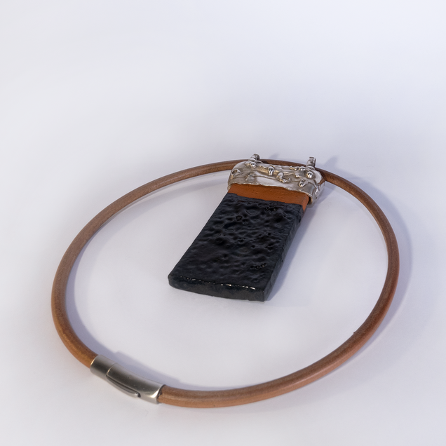 Pendant made of a rectangular black glazed ceramic piece with a silver cap, part of the ceramic is natural reddish, it hangs on a heavy reddish leather strap; the glaze "Fat Lava" looks like the surface of the moon 