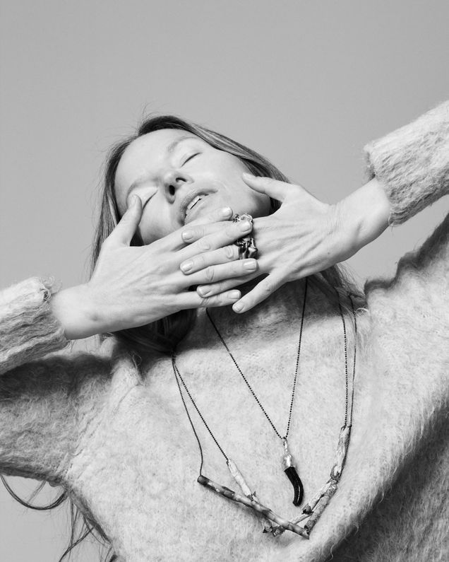 Artist Michaela Schwarz-Weismann wears three necklaces and two rings by Ela Nord over a knitted sweater, she clasps her hands under her chin, her eyes are closed