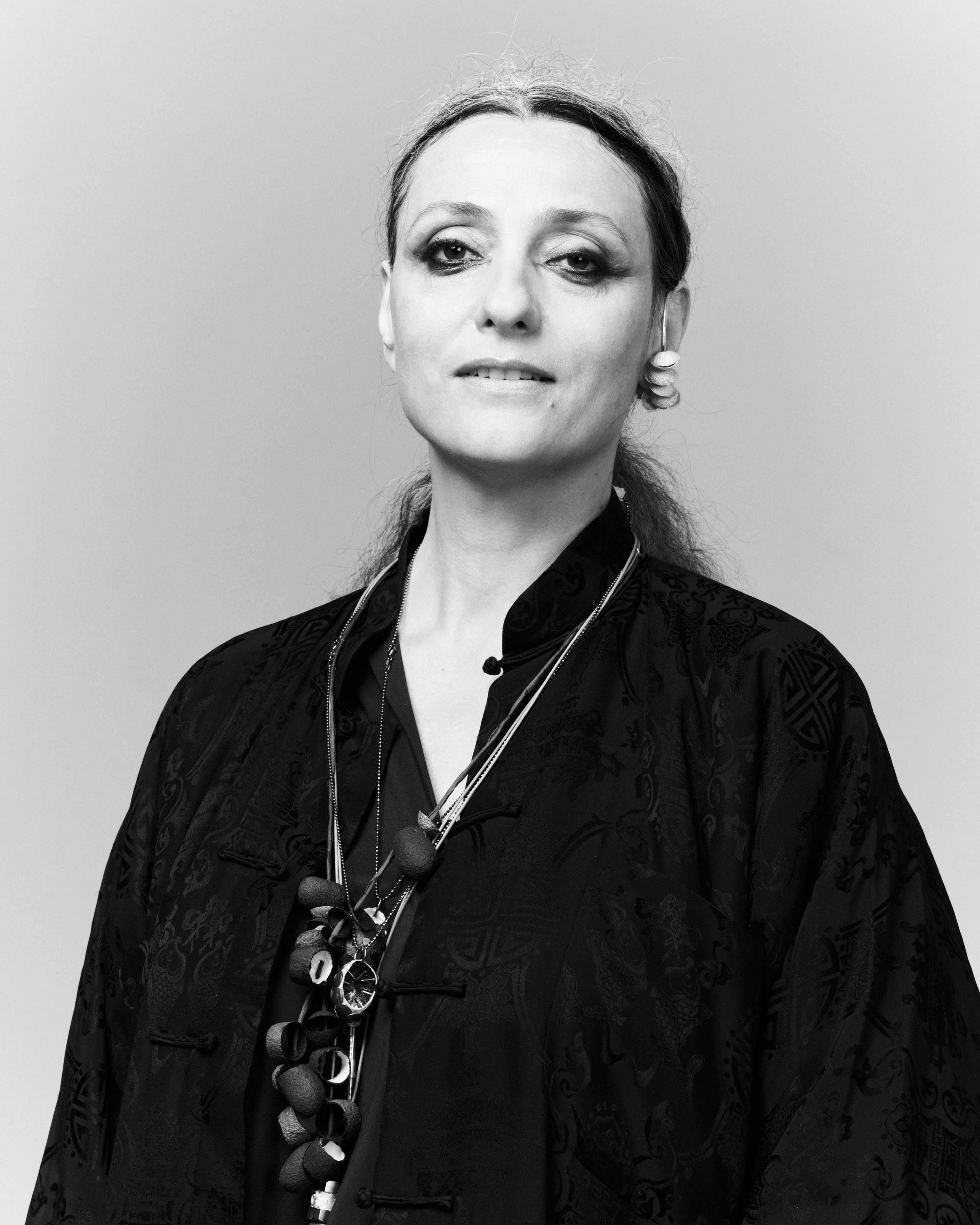 Künstlerin Maja Vukoje trägt mehrere Seidenkokon-Halsketten über einer schwarzen Bluse, ein Ohrring mit weißen Kokonteilen hängt an ihrem Ohr, sie schaut ernst, ihr langes Haar ist hochgebunden