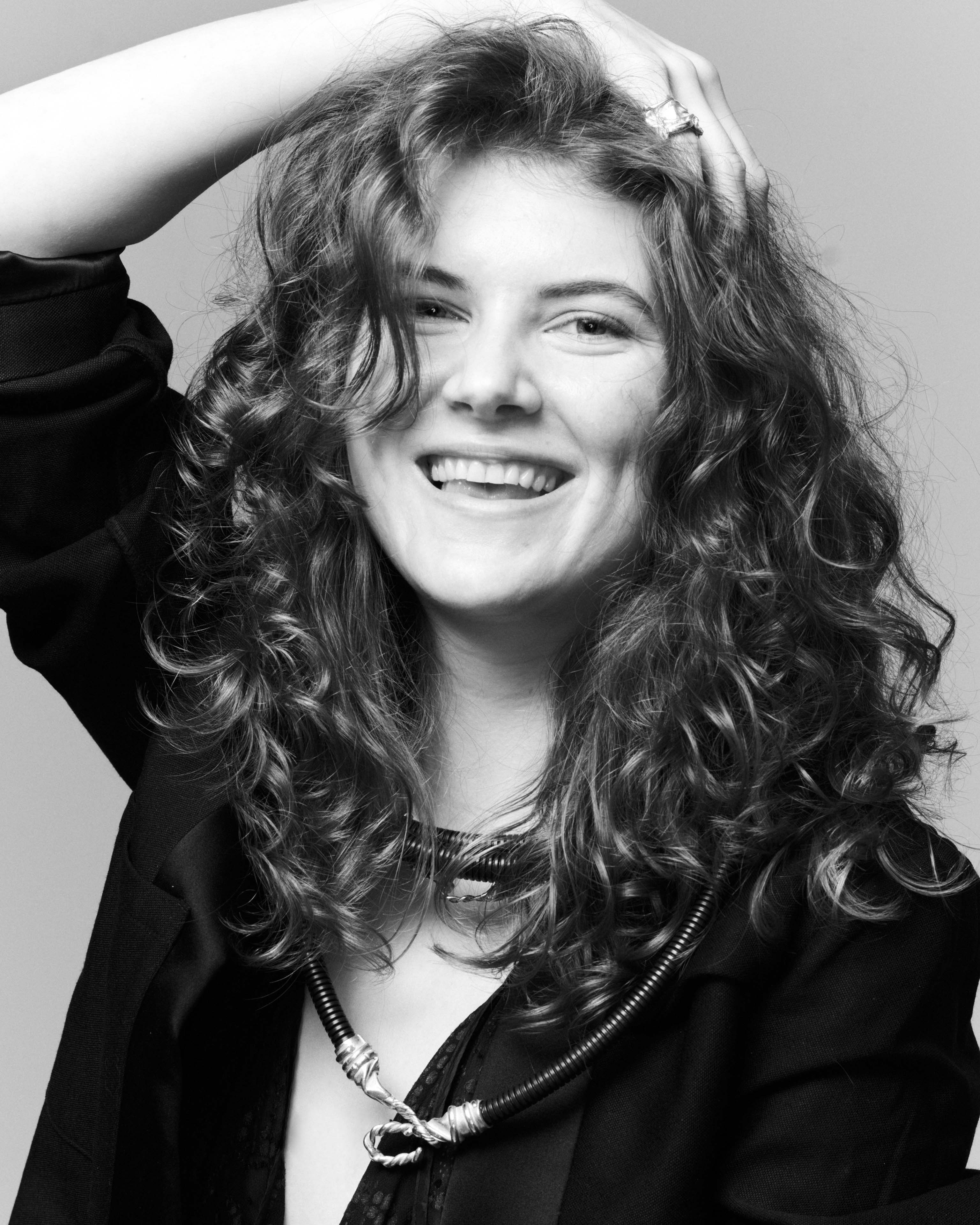 Young woman wears three hose chains over a black blazer, she laughs and has her hand on her head, she has long curly hair