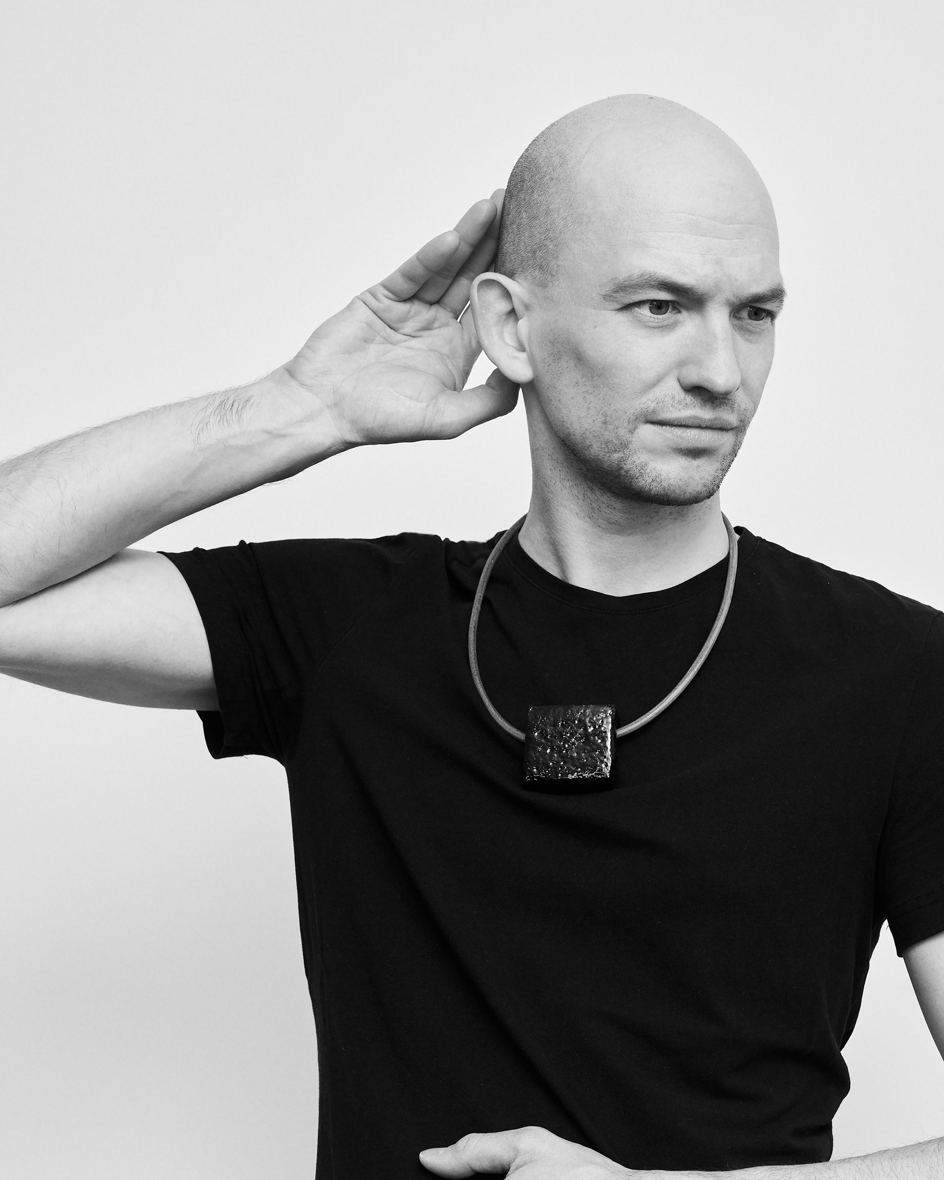 Artist Nick Oberthaler wears a short necklace with a silver ceramic pendant on a leather strap over a black T-shirt, his look is serious